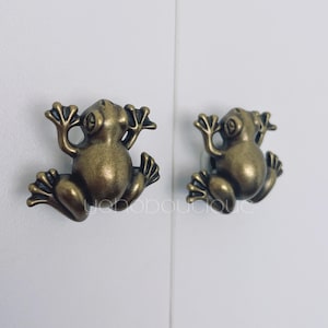 May include: Two bronze-colored frog-shaped cabinet knobs. The knobs are detailed with textured skin and outstretched limbs, giving them a whimsical appearance. They are mounted on a white surface, suggesting they are ready for installation on furniture.