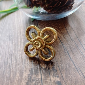 Könnte beinhalten: Goldfarbener Metall-Schrankknopf mit einem dekorativen Rosenmuster. Der Knopf hat die Form eines Kleeblatts.