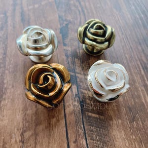 May include: Four rose-shaped cabinet knobs in different finishes: gold, silver, white, and a darker gold. The knobs are on a wooden surface.