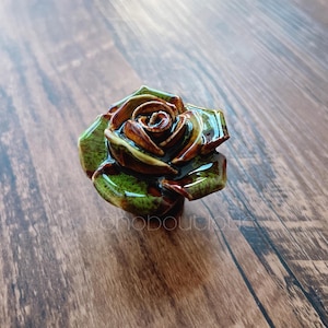 May include: A ceramic rose with green and brown hues. The handcrafted flower has detailed petals and a glossy finish. The rose is a decorative item, possibly a cabinet knob or furniture accent.