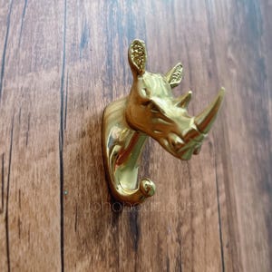 Könnte beinhalten: Ein goldfarbener Wandhaken in Form eines Nashornkopfes. Der Haken ist auf einer Holzoberfläche montiert. Der Nashornkopf ist mit einem Horn und Ohren detailliert. Der Haken ist unten gebogen.