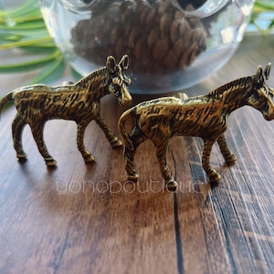 May include: Two bronze-coloured miniature horse figurines. The horses have textured manes and bodies, standing on four legs. The figurines are on a wooden surface, with a glass container and greenery in the background. The text "yohoboutique" is visible.