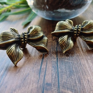 Może przedstawiać: Dwa antyczne, złote gałki do szafek w kształcie kokardki. Gałki mają szczegółowe, teksturowane powierzchnie z pionowymi liniami i centralnym akcentem z koralików. Gałki leżą na ciemnej drewnianej powierzchni z rozmytym tłem.