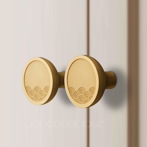 Pode incluir: Dois botões redondos de ouro para armário com um design de ondas. Os botões estão presos a uma porta branca.