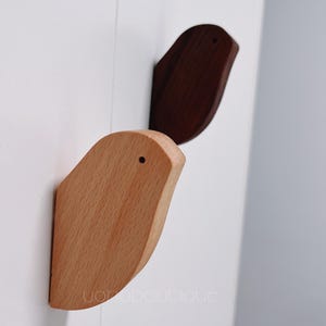 Op de afbeelding: Twee houten vogelvormige wandhaken, een in lichtbruin en een in donkerbruin. De haken zijn bevestigd aan een witte muur.