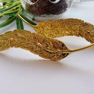 Könnte beinhalten: Zwei goldfarbene, metallene Dekorationsstücke in Federform mit filigranen Details. Die Federn haben eine strukturierte Oberfläche und sind gebogen.