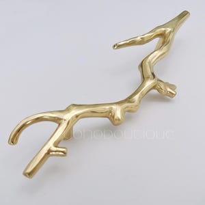Op de afbeelding: Gouden metalen kastgreep in de vorm van een gestileerde tak. De greep heeft een gladde, gepolijste afwerking.