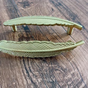 Könnte beinhalten: Zwei goldfarbene, federförmige Schrankgriffe. Die Griffe sind aus Metall und haben ein detailliertes Federdesign.