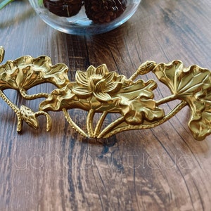 Op de afbeelding: Een gouden metalen lotusbloemsculptuur met gedetailleerde bladeren en een centrale bloem. De sculptuur is geplaatst op een houten oppervlak, met een glazen kom met decoratieve items op de achtergrond.