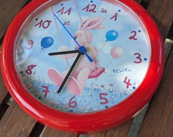 May include: Red round clock with a white face and black numbers. A cartoon bunny with blue eyes is on the clock face, holding onto a swing with blue and pink balloons. The clock face says "Felix..." at the bottom.
