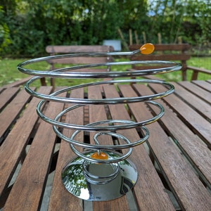 May include: A chrome metal fruit bowl shaped like a spiral with two orange accents. The bowl is sitting on a wooden table.
