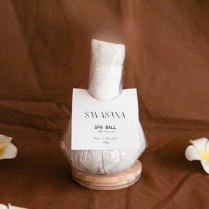 Puede incluir: Una bola de spa con un mango cilíndrico blanco y un cuerpo redondo gris, presentada sobre una base de madera. La etiqueta dice "SAVASANA SPA BALL 100% Natural" y el peso es de 150g. La bola de spa está envuelta en plástico transparente.
