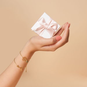 May include: A hand holding a small white gift box with a pink ribbon and bow. The box is labeled "Merci" in white lettering. The hand is wearing two gold bracelets, one with a lotus flower charm and the other with a "Love" charm.
