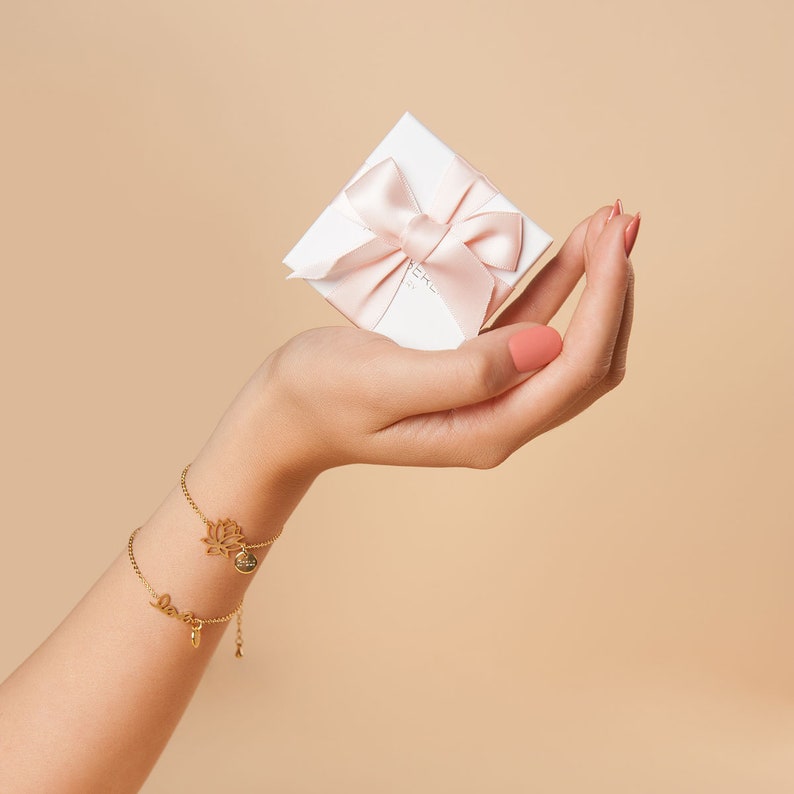 May include: A hand holding a small white gift box with a pink ribbon and bow. The box is labeled "Merci" in white lettering. The hand is wearing two gold bracelets, one with a lotus flower charm and the other with a "Love" charm.