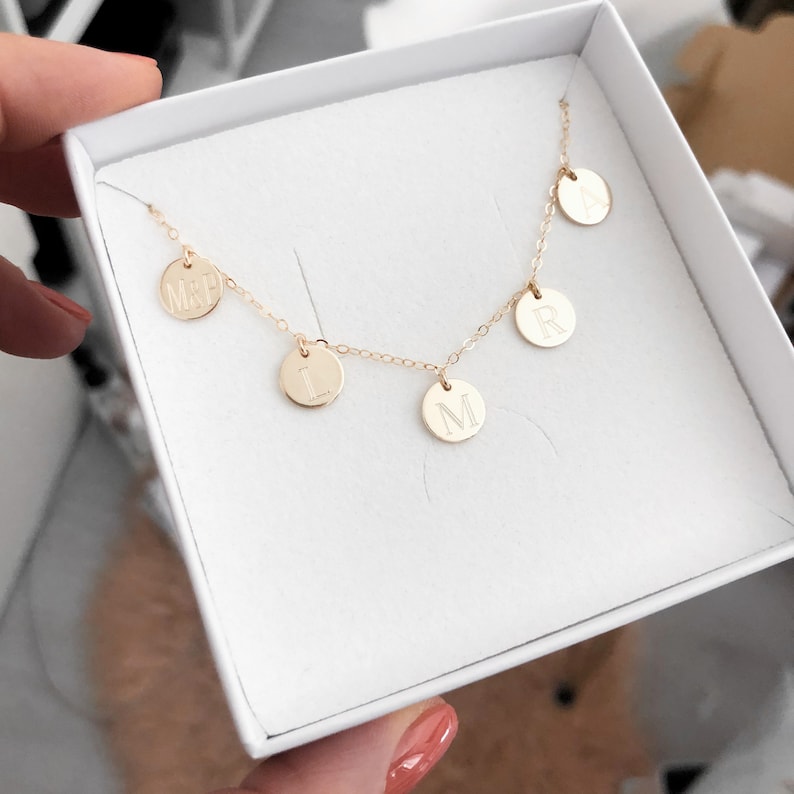 May include: A gold necklace with five small round charms, each with a letter engraved on it. The letters spell out "MARIL." The necklace is displayed in a white box.