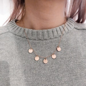 May include: A delicate gold necklace with five small, round pendants hanging from it. The necklace is worn by a person wearing a light grey sweater.