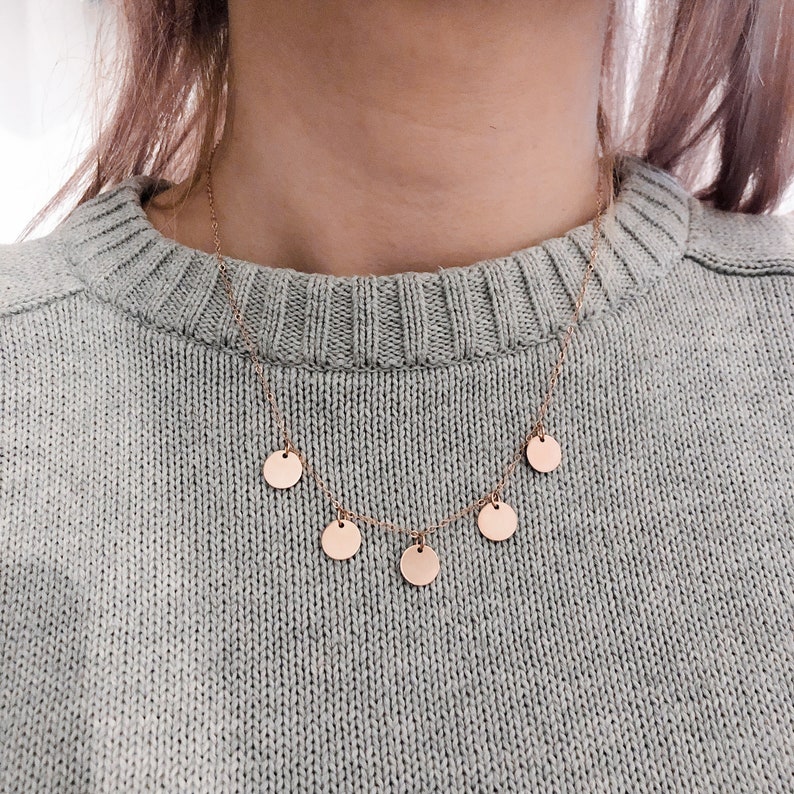 May include: A delicate gold necklace with five small, round pendants hanging from it. The necklace is worn by a person wearing a light grey sweater.