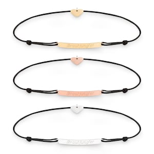 May include: Three black string bracelets with a small heart charm and a metal plate with the inscription "Brautjungfer" in gold, rose gold, and silver.