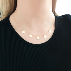 May include: A gold necklace with five small, round, pendant charms. The necklace is on a model wearing a black shirt.