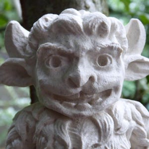 May include: A detailed, off-white stone sculpture of a mythical creature with a smiling face, large ears, and horns. The sculpture has a textured surface and is set against a blurred green background.