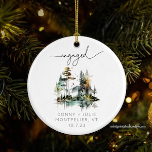 May include: White ceramic Christmas ornament with a watercolor painting of a forest scene and the words "engaged" and "Donny + Julie Montpelier, VT 10.7.23".