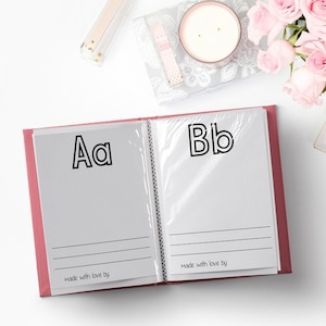 May include: An open pink scrapbook with white pages featuring the letters "Aa" and "Bb" in black outlined font. The pages have lines for writing. The scrapbook is on a white surface with pink roses, candles, and a white dish with the text "hello gorgeous" in gold lettering.