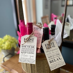 May include: Three wine bottles with gift tags attached. The tags are for a first anniversary, a first house, and a first Valentine's Day. The bottles are in a metal bucket with pink tissue paper.