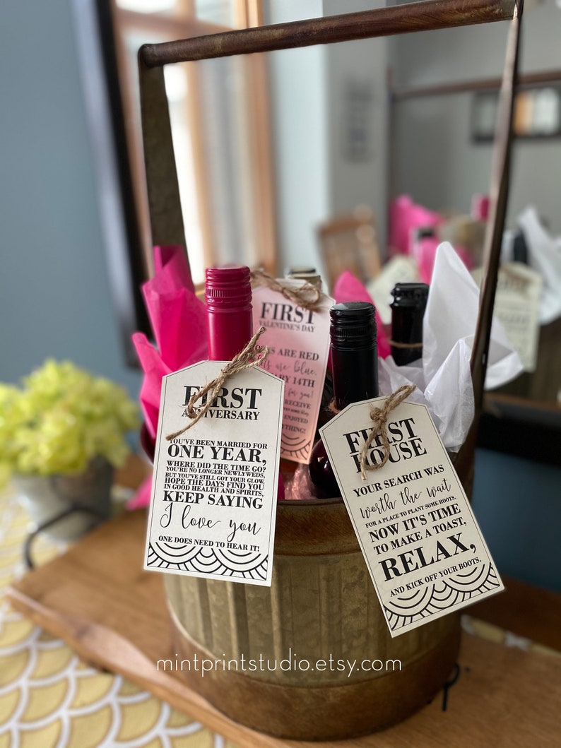 May include: Three wine bottles with gift tags attached. The tags are for a first anniversary, a first house, and a first Valentine's Day. The bottles are in a metal bucket with pink tissue paper.