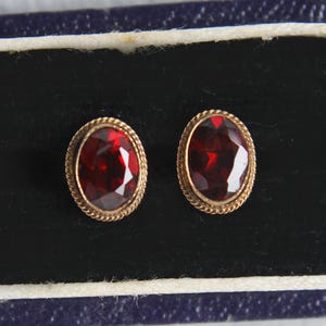 Op de afbeelding: Een paar ovale oorstekers met rode edelstenen en goudkleurige, touwtextuur randen. De oorbellen worden gepresenteerd in een donkerblauwe doos met een zwart interieur.