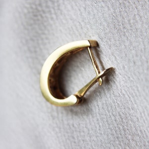 K&ouml;nnte beinhalten: Ein einzelner goldfarbener Creolen-Ohrring mit einem Klappverschluss. Der Ohrring hat ein abgerundetes, D-f&ouml;rmiges Design. Der Hintergrund ist ein hellgrauer Stoff.