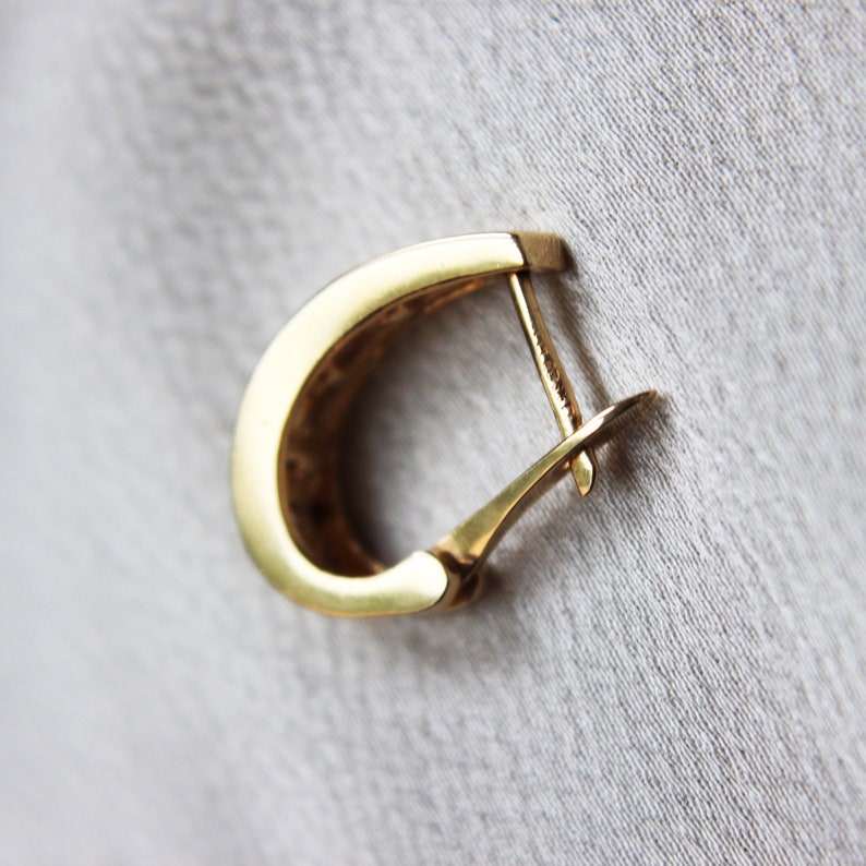 K&ouml;nnte beinhalten: Ein einzelner goldfarbener Creolen-Ohrring mit einem Klappverschluss. Der Ohrring hat ein abgerundetes, D-f&ouml;rmiges Design. Der Hintergrund ist ein hellgrauer Stoff.