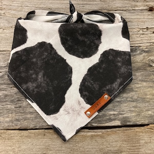 Peut inclure: Un bandana blanc et noir &agrave; imprim&eacute; vache avec une &eacute;tiquette en cuir marron sur laquelle est inscrit "DUKE".