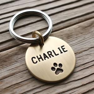May include: A gold-colored round dog tag with a silver key ring. The tag has the name "CHARLIE" engraved in black lettering and a paw print.