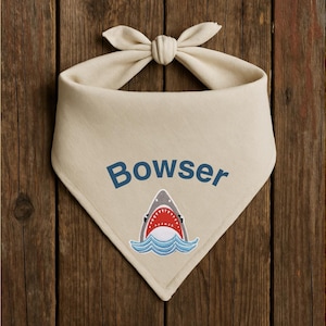 May include: A beige dog bandana with a blue shark graphic and the name "Bowser" embroidered in blue.