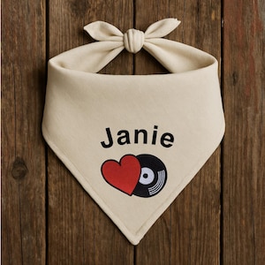 Könnte beinhalten: Cremefarbenes Hundehalstuch mit dem Namen "Janie" in Schwarz, einem roten Herz und einer schwarzen Schallplatten-Grafik. Das Halstuch ist oben mit einer Schleife gebunden.