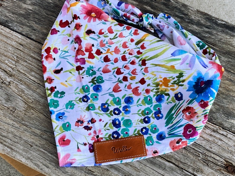 May include: A white bandana with a floral print featuring colorful flowers and green leaves. The bandana has a brown leather tag with the word "Willow" printed on it.