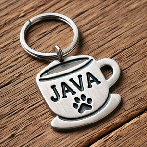 Könnte beinhalten: Silberner Schlüsselanhänger mit einem Kaffeetassen-Anhänger. Der Anhänger hat das Wort "JAVA" und einen Pfotenabdruck.