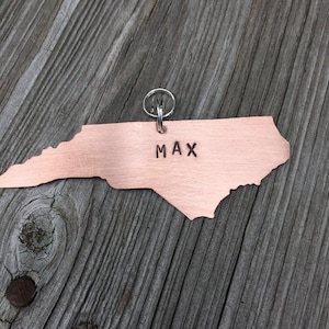 K&ouml;nnte beinhalten: Ein kupferfarbener Anh&auml;nger in Form des Bundesstaates North Carolina mit einem silbernen Schl&uuml;sselring. Der Anh&auml;nger tr&auml;gt den eingravierten Namen "MAX".