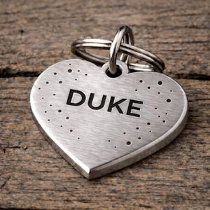 May include: A heart-shaped, brushed metal pet tag with the name "DUKE" in bold, black letters. The tag has small, evenly spaced dot details and a silver ring for attachment. The tag is shown on a wooden surface.