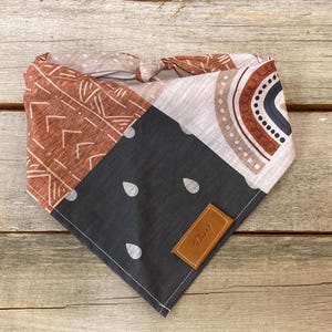 Peut inclure: Un bandana triangulaire avec un motif patchwork composé de motifs géométriques, un arc-en-ciel et des gouttes de pluie. Le bandana est en tissu de coton et possède une étiquette en cuir marron avec le mot "Dog" dessus.
