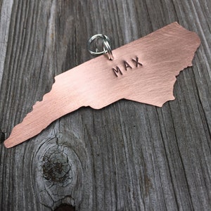 K&ouml;nnte beinhalten: Ein kupferner Hundemarken in Form des Bundesstaates North Carolina, mit dem Namen Max eingraviert. Die Marke hat einen silbernen Ring.