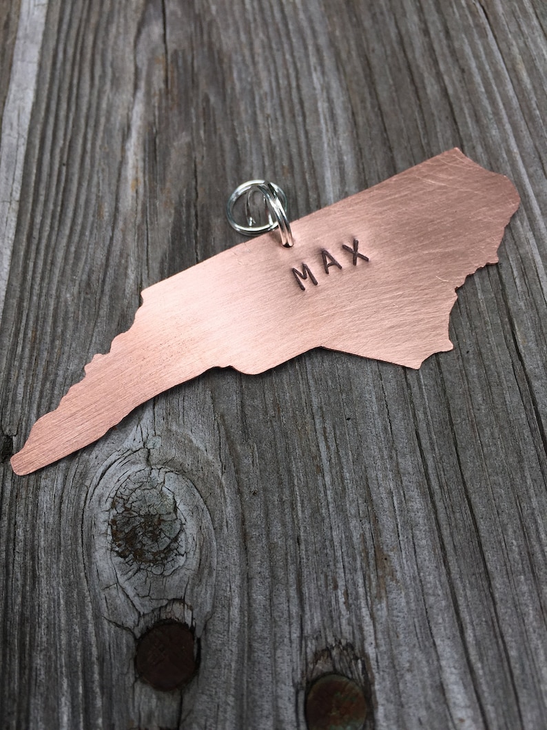 K&ouml;nnte beinhalten: Ein kupferner Hundemarken in Form des Bundesstaates North Carolina, mit dem Namen Max eingraviert. Die Marke hat einen silbernen Ring.
