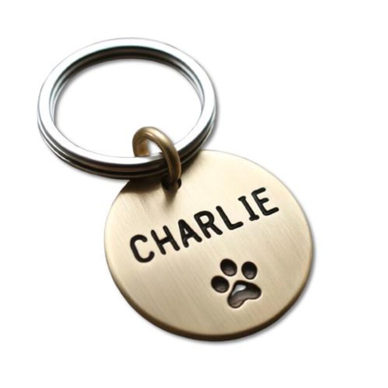 May include: Gold-toned round dog tag with a silver key ring. The tag has the name "CHARLIE" engraved in black lettering and a paw print.
