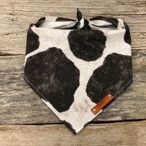Peut inclure: Un bandana blanc et noir &agrave; imprim&eacute; vache avec une &eacute;tiquette en cuir marron qui dit "DUKE".