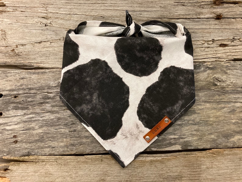 Peut inclure: Un bandana blanc et noir &agrave; imprim&eacute; vache avec une &eacute;tiquette en cuir marron qui dit "DUKE".