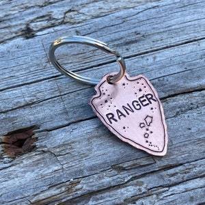 May include: A copper-coloured arrowhead keyring with the word "RANGER" engraved on it. The keyring is attached to a silver key ring.
