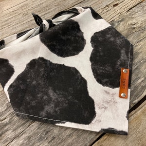 Peut inclure: Un bandana blanc et noir &agrave; imprim&eacute; vache avec une &eacute;tiquette en cuir marron sur laquelle est inscrit "DUKE".