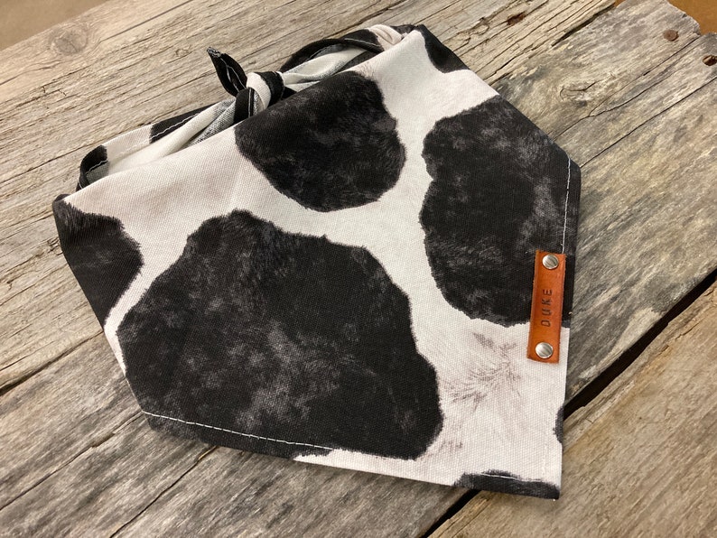 Peut inclure: Un bandana blanc et noir &agrave; imprim&eacute; vache avec une &eacute;tiquette en cuir marron sur laquelle est inscrit "DUKE".