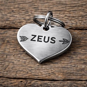 Könnte beinhalten: Ein herzförmiger, silberfarbener Haustieranhänger mit dem Namen "ZEUS" in Schwarz graviert, umrahmt von Pfeildesigns. Der Anhänger ist an einem silbernen Ring befestigt. Der Hintergrund ist eine rustikale Holzoberfläche.
