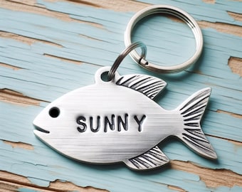 May include: Silver fish-shaped key chain with the name "SUNNY" engraved on it. The keychain is attached to a silver ring.
